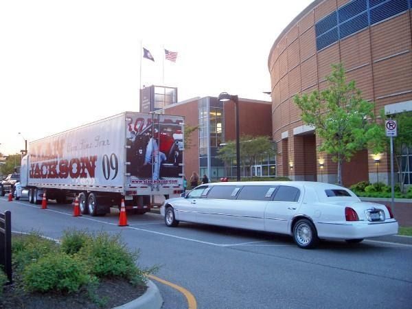White stretch limousine parked beside a promotional truck outside a brick arena at dusk
