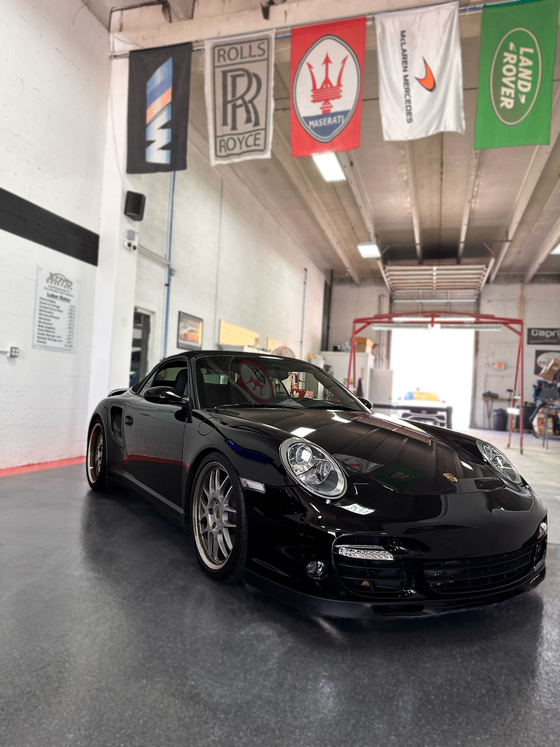 Black Porsche convertible in a garage with luxury car flags hanging above.