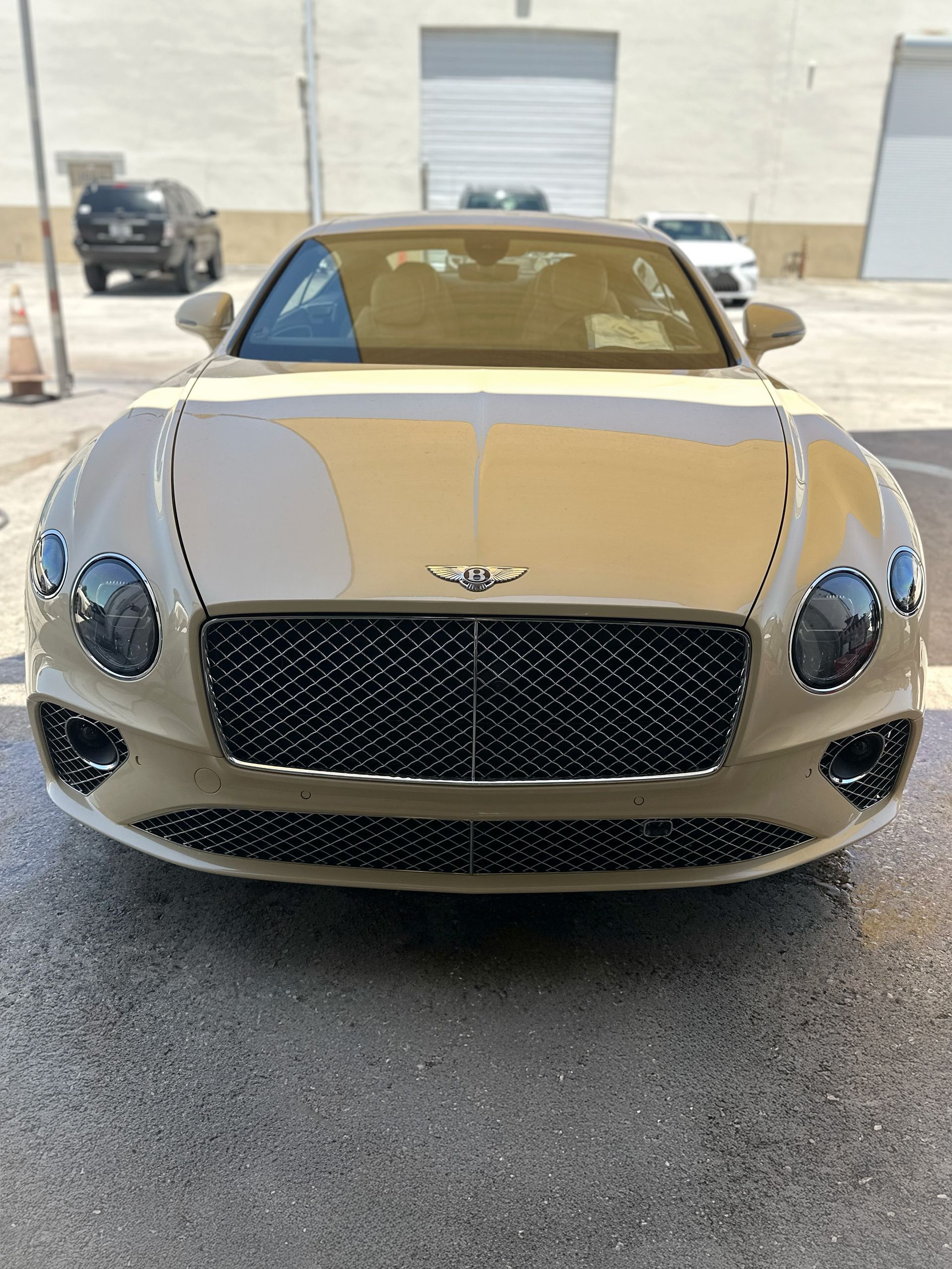 Tan Bentley car parked in front of a building with other vehicles in the background.