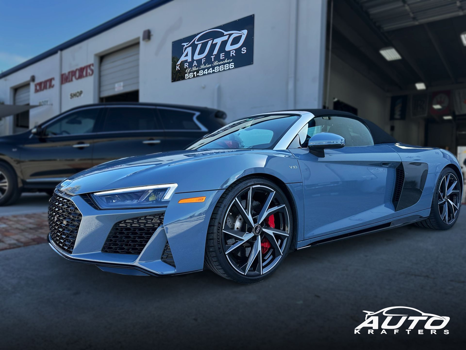 Blue Audi R8 Spyder convertible parked in front of a car repair shop, with a black SUV in the background.