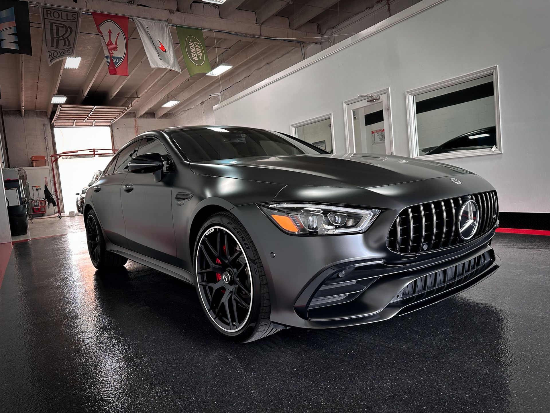Matte gray Mercedes-AMG GT 4-door coupe parked in a garage with a glossy floor and flags overhead.