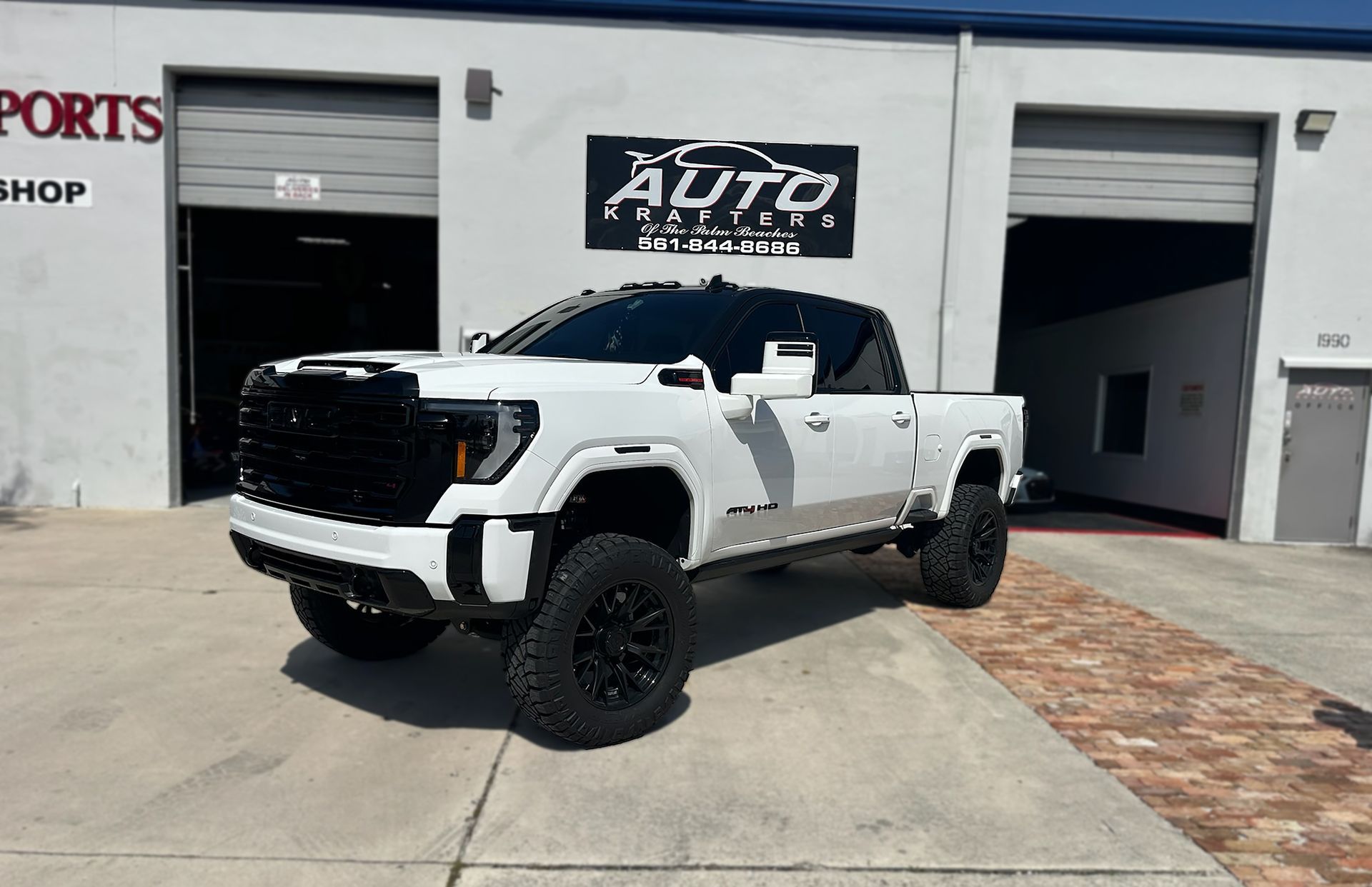 White lifted GMC truck parked in front of a auto shop. Black rims and grill, gray building.
