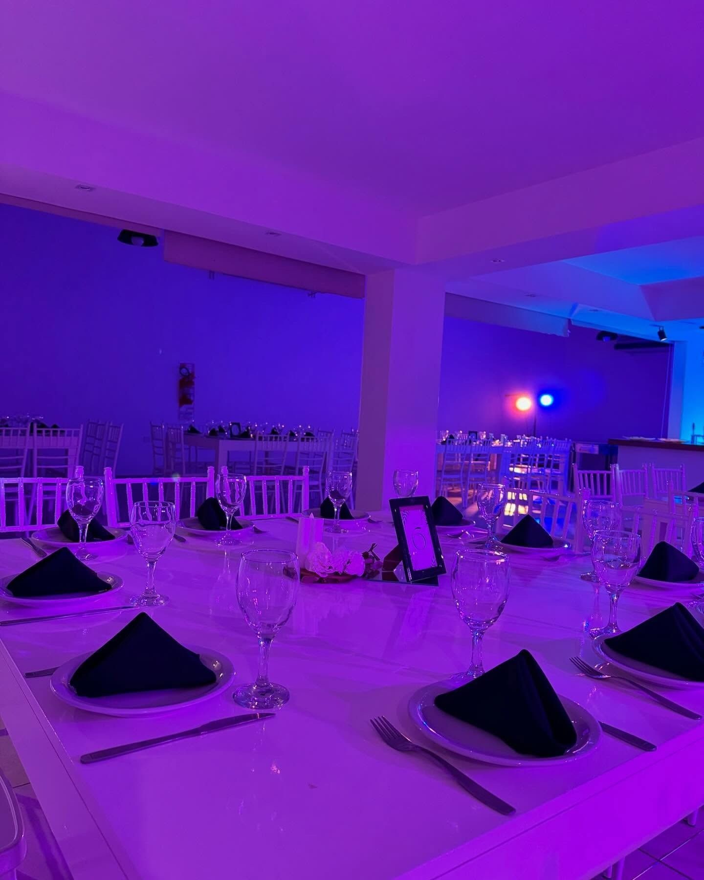 Juego de comedor formal para fiesta con mesas blancas, iluminación violeta y servilletas negras.
