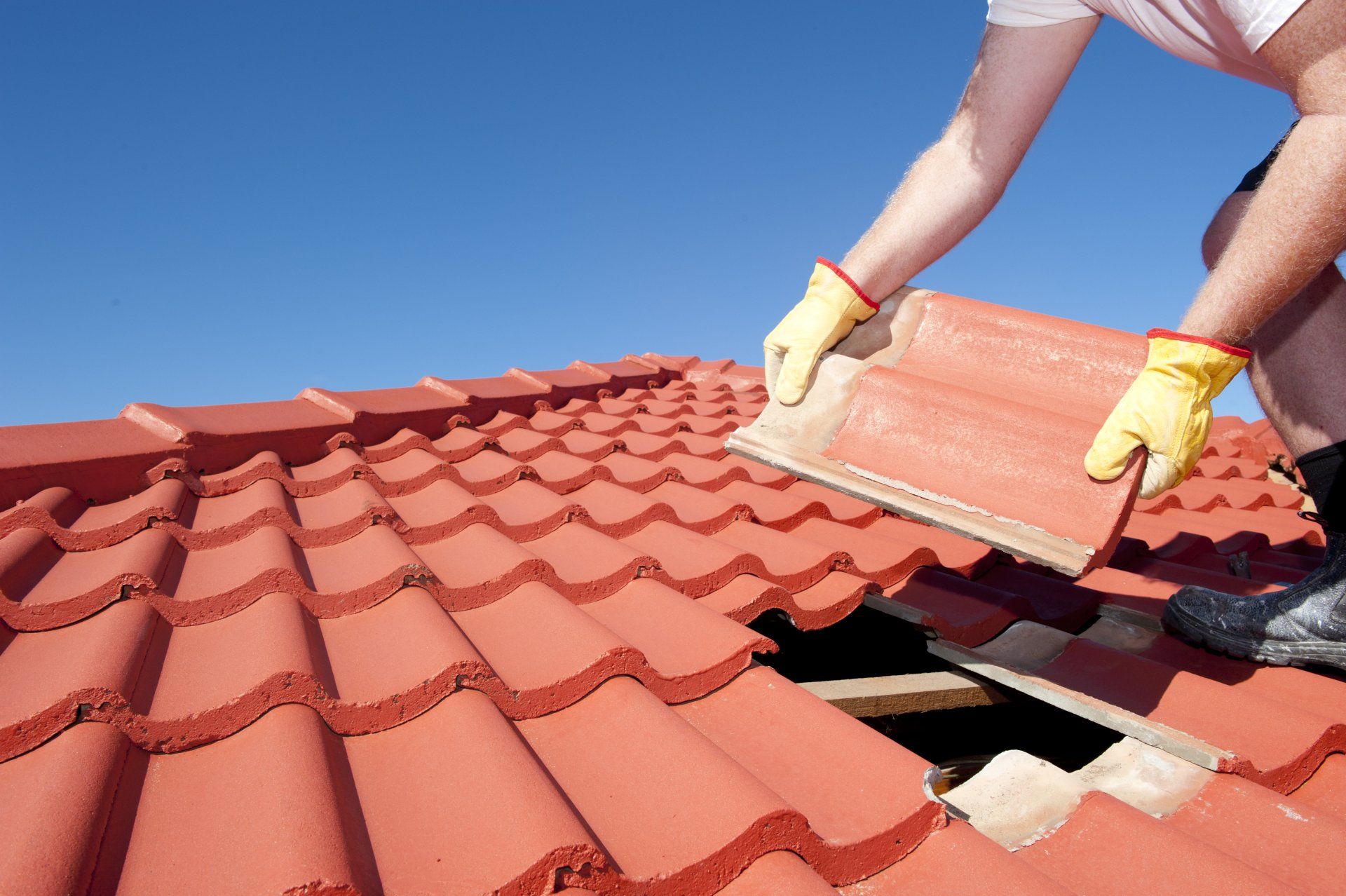 Roof repair, worker with yellow gloves replacing red tiles or shingles on house with blue sky as background and copy space