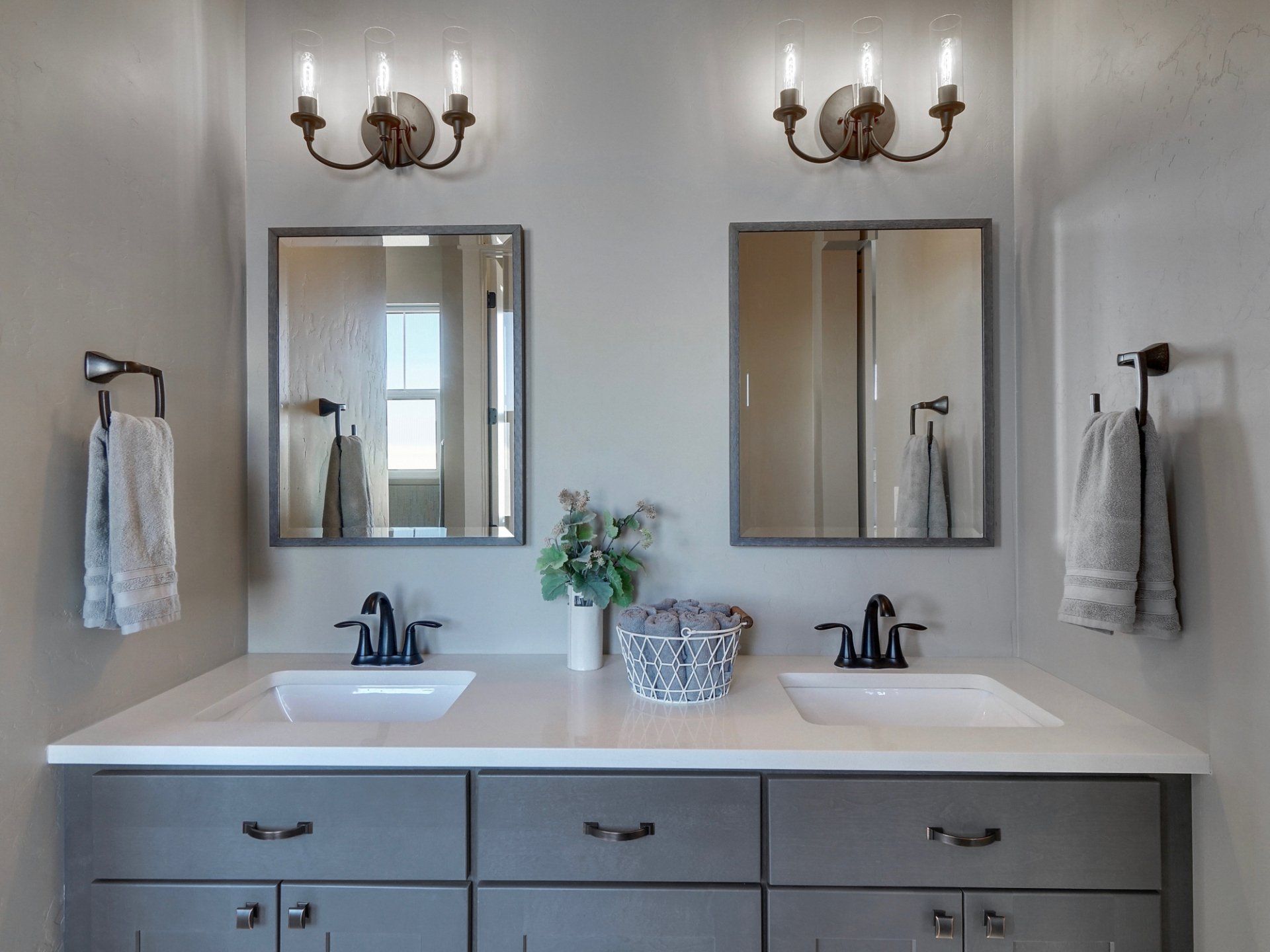 Bathroom with two sinks, gray cabinets, mirrors, and sconces. Towels hang on bars.