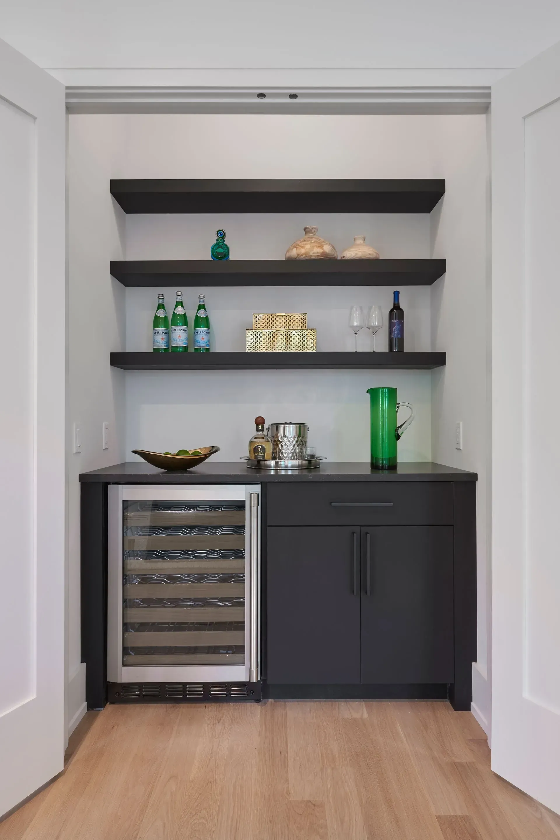 Built-in bar with black shelving, countertop, cabinet and wine fridge; bottles and decor displayed.