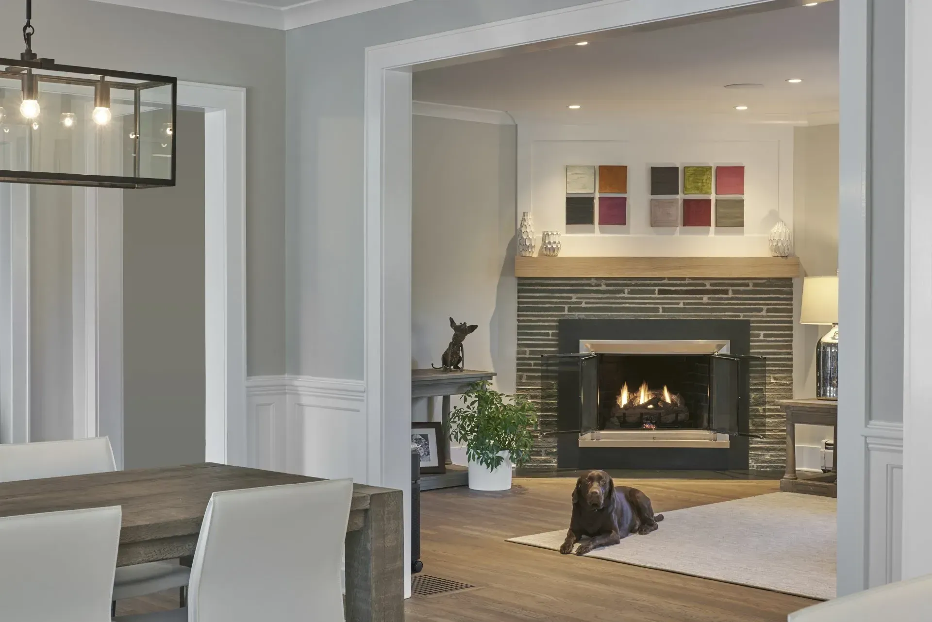 Dining room view into living room with dog; fireplace, art, modern lighting, and wood flooring.