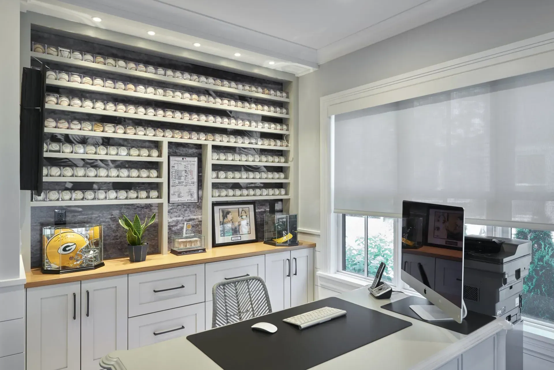 Home office with built-in shelves displaying baseballs. Desk with computer, printer, and a window with a blind.