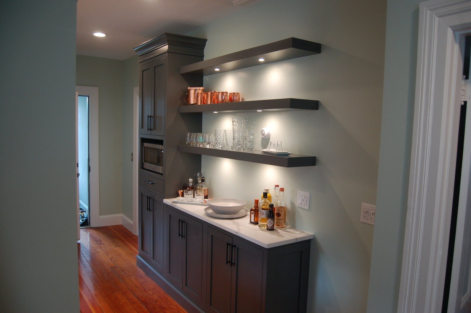 Gray built-in bar with shelves, under-cabinet lighting, and white countertop. Copper mugs and glassware on shelves.