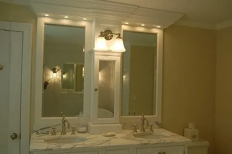 Bathroom vanity with two mirrors, a central cabinet, and sinks. White cabinetry and light-colored countertop.