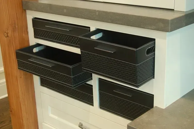Black storage bins pull out from a white cabinet under a gray countertop.