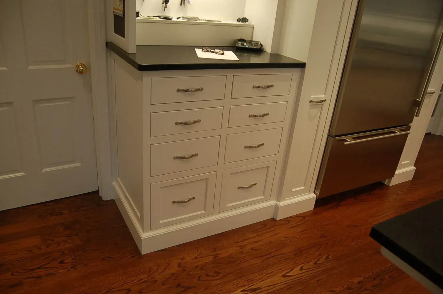 White cabinet with eight drawers, black countertop, stainless steel refrigerator, wooden floor.