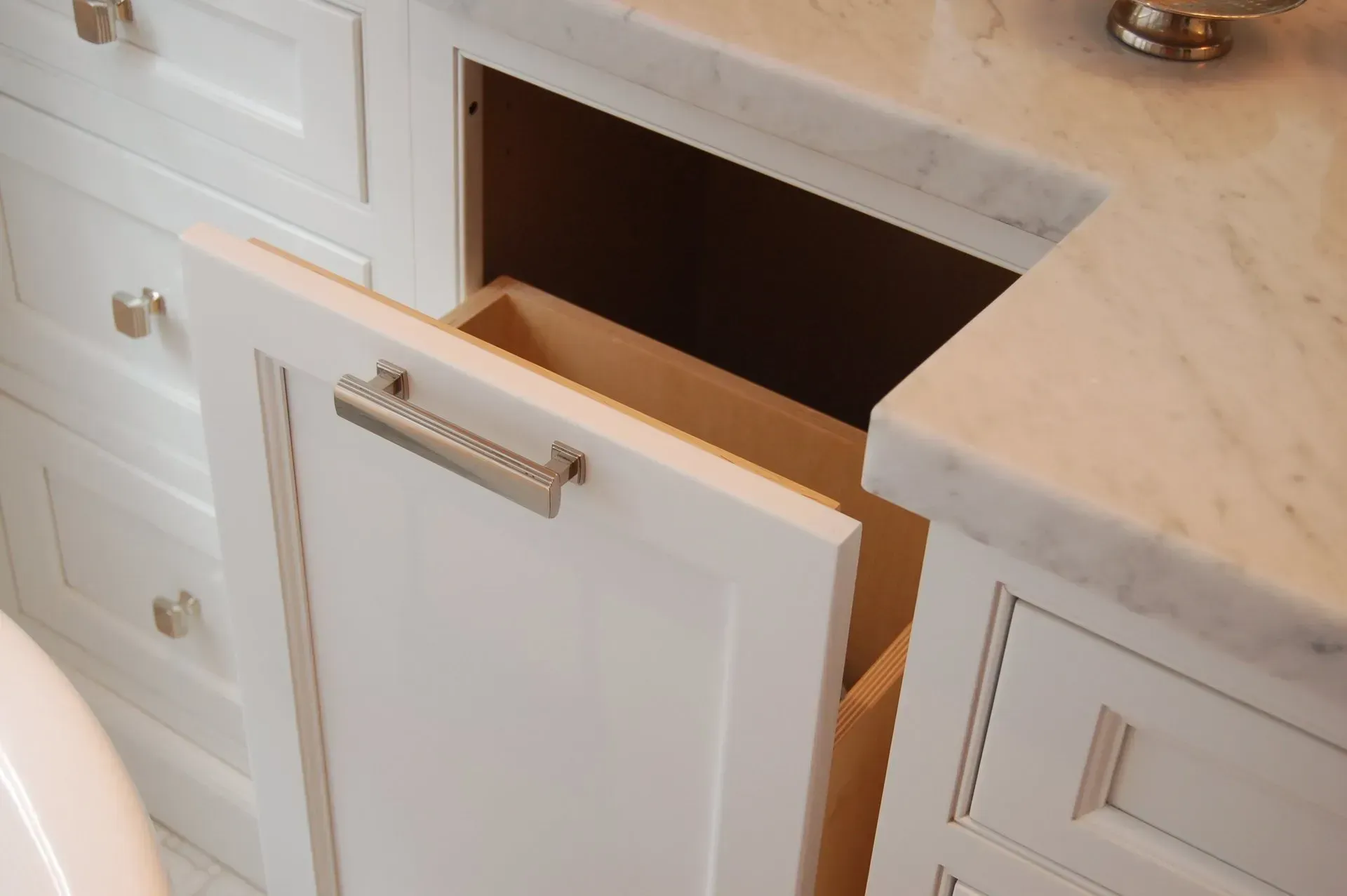 White bathroom vanity with open pull-out trash can. Marble countertop, silver hardware.