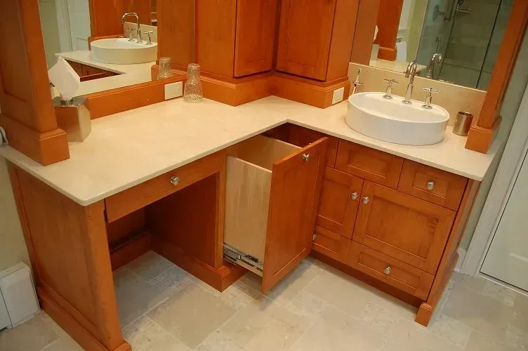 Corner bathroom vanity with open trash can. Wooden cabinets and beige countertop. White sink and mirror.