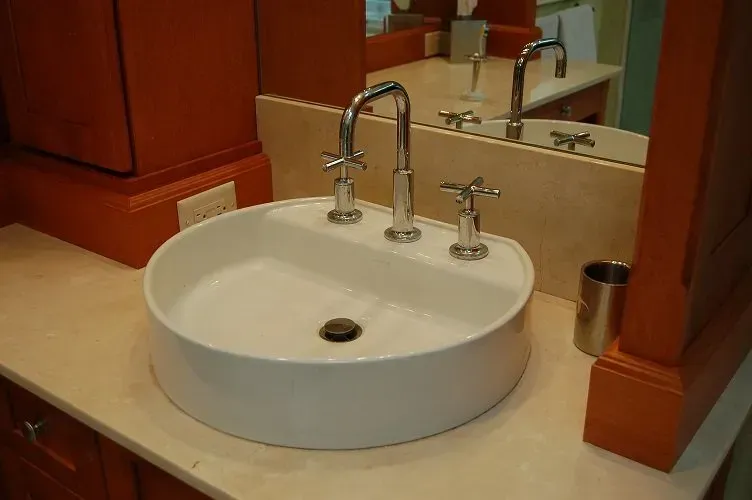 White, round bathroom sink with chrome faucet and orange cabinets, on a light beige countertop.