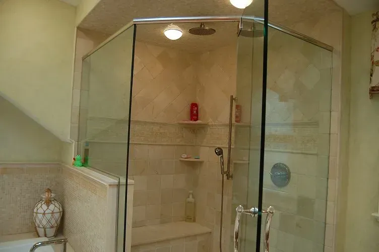 Shower with glass doors and beige tile, built-in bench, and shower head, in a bathroom setting.