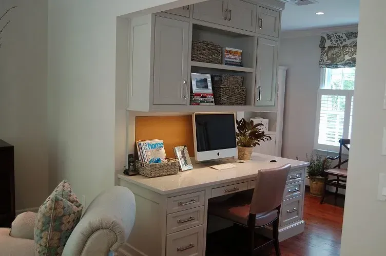 Home office with built-in desk, cabinets, computer, and chair. Light-colored walls, wood floor, and natural light.