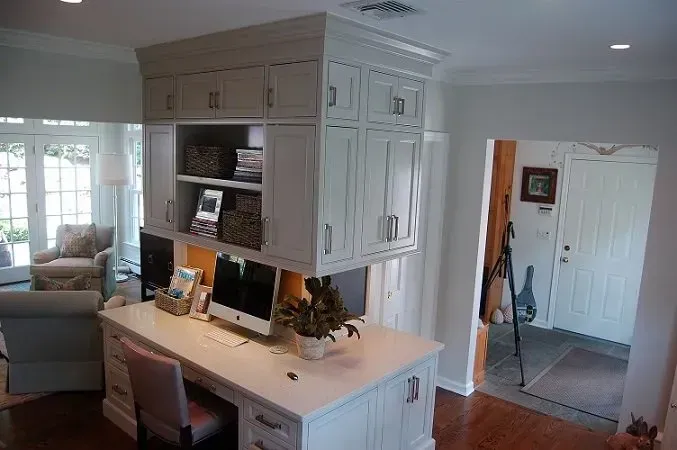 White built-in desk with overhead cabinets; two monitors, chair, entry door, window to seating area.