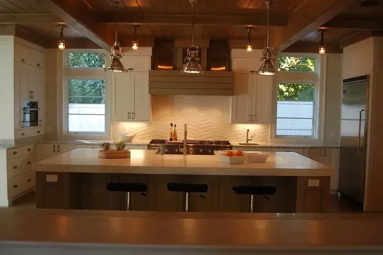Modern kitchen with large island, stove, white cabinets, light wood ceiling, and pendant lights.