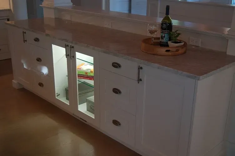 White kitchen island with countertop, cabinets, and glass-door refrigerator. Bottle, glass, and tray on countertop.