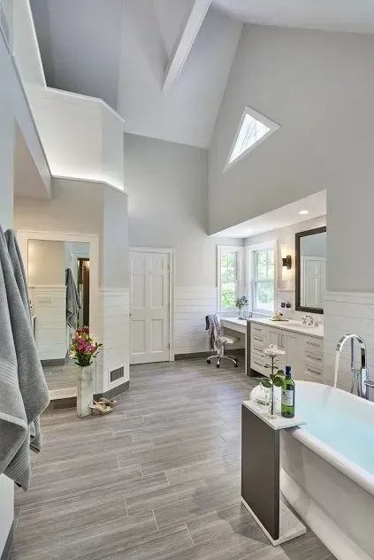Spacious, light gray bathroom with wood-look tile floor, a soaking tub, and vaulted ceiling with a triangular window.