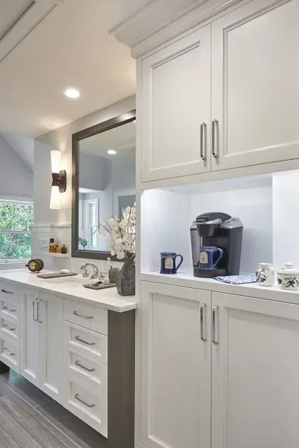 White kitchen cabinets with a coffee station. Cabinets have silver handles. Bright overhead lighting.