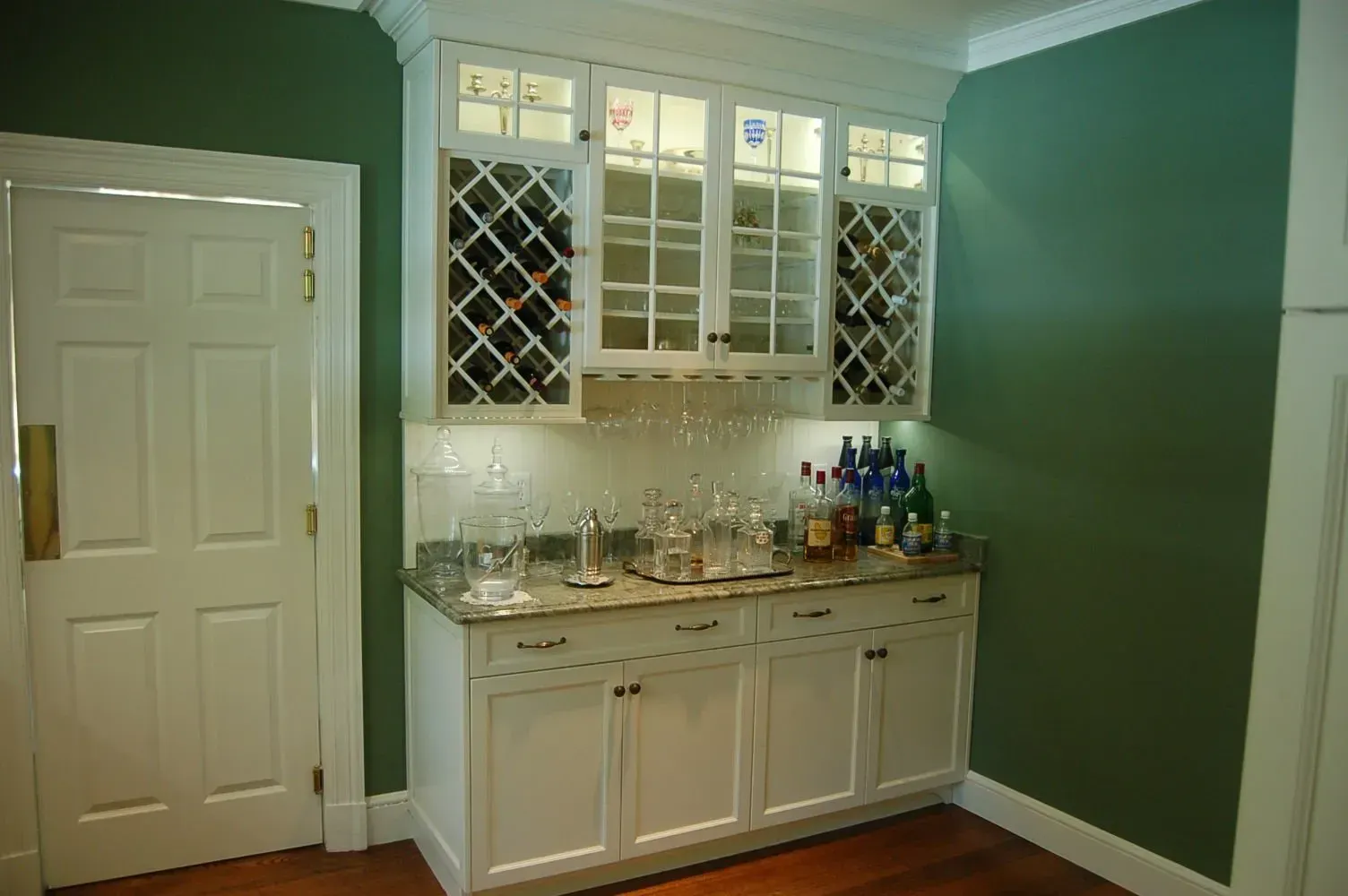 White cabinet bar with wine racks, glass shelves, granite countertop, and glassware against a green wall.