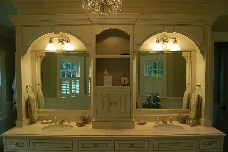Bathroom vanity with two arched mirrors, matching sconces, and a central cabinet; cream-colored, ornate woodwork.