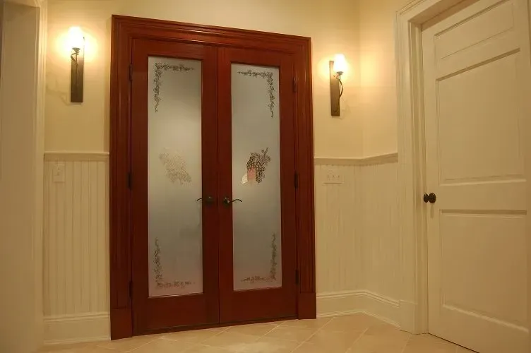 Double red doors with frosted glass panels, flanked by sconces, next to a white door in a cream-colored hallway.