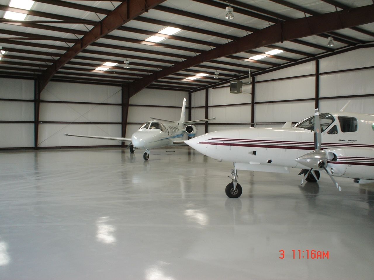 Two small planes are parked in a hangar at 11:16 am