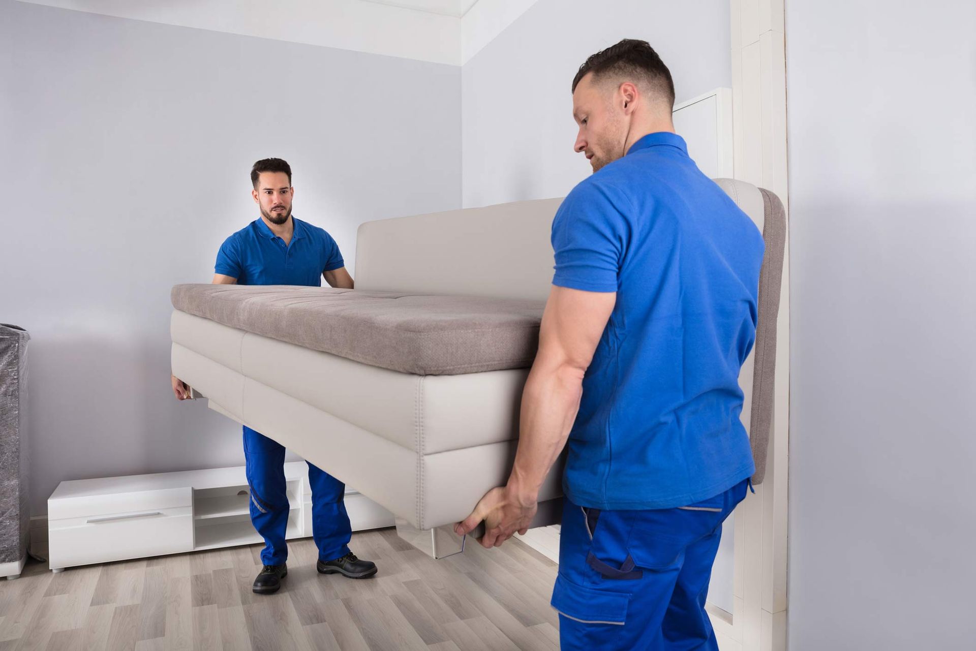 Two local furniture removalists, in blue uniforms, are holding a sofa in the living room.
