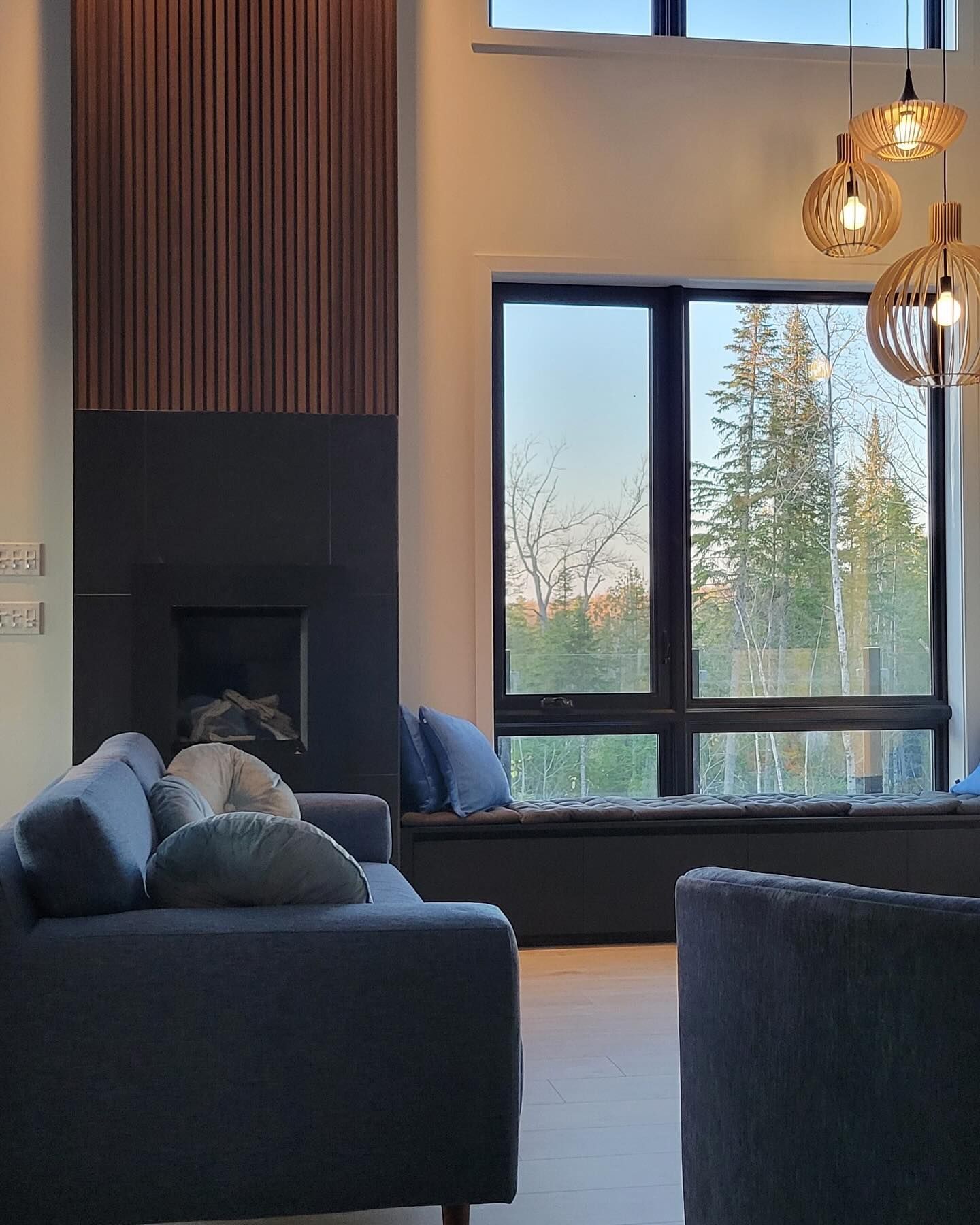 Séjour avec grande fenêtre donnant sur la forêt, canapé foncé, luminaires suspendus.
