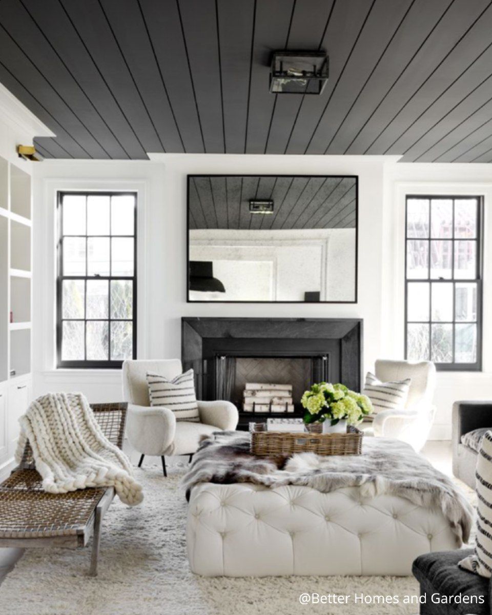 Salon avec cheminée noire, plafond sombre, grand miroir, chaises blanches et pouf capitonné.