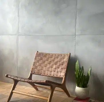 Une chaise en cuir tressé repose contre un mur gris texturé, avec une plante en pot sur un plancher en bois.