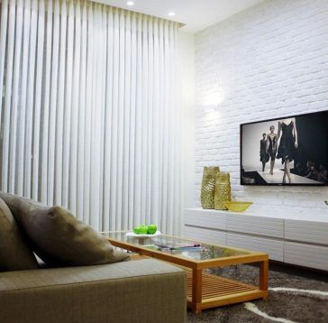 Salon confortable avec rideaux blancs, mur de briques, télévision et canapé ; une table basse en verre avec des pommes vertes.