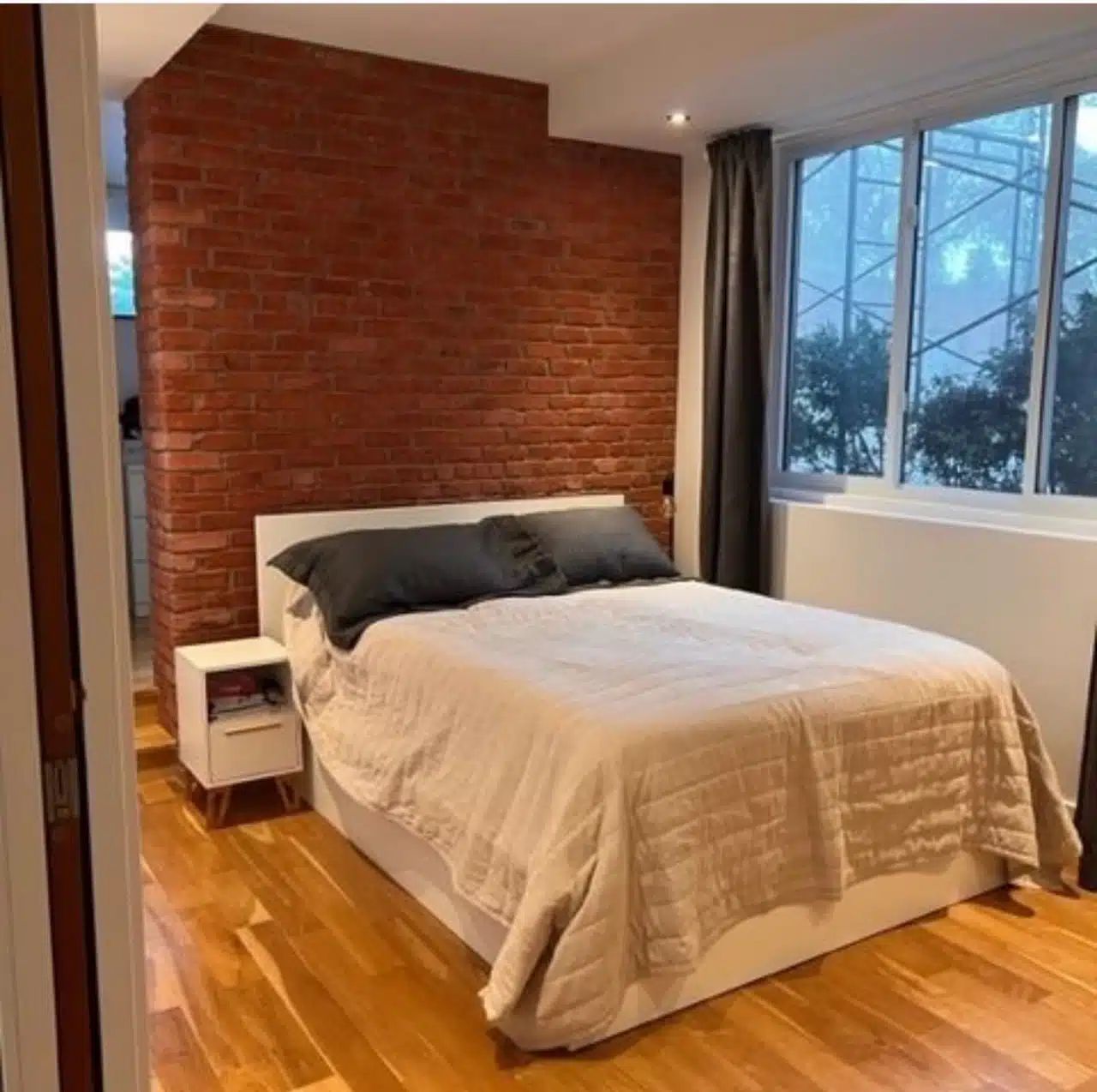 Chambre avec mur en briques apparentes, lit blanc, table de chevet et grande fenêtre avec vue sur la ville.