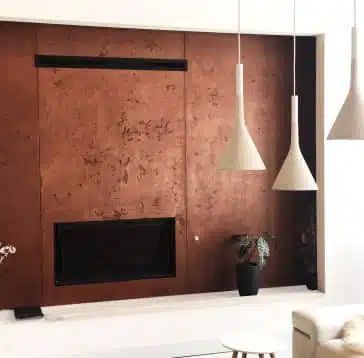 Mur aux tons cuivrés avec cheminée et évent rectangulaire, lumières blanches suspendues en forme de cône et plante en pot dans un cadre moderne.