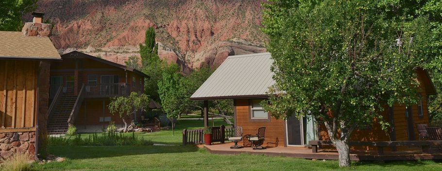 Rustic cabins and a small house in a green yard with trees, backed by red rock cliffs