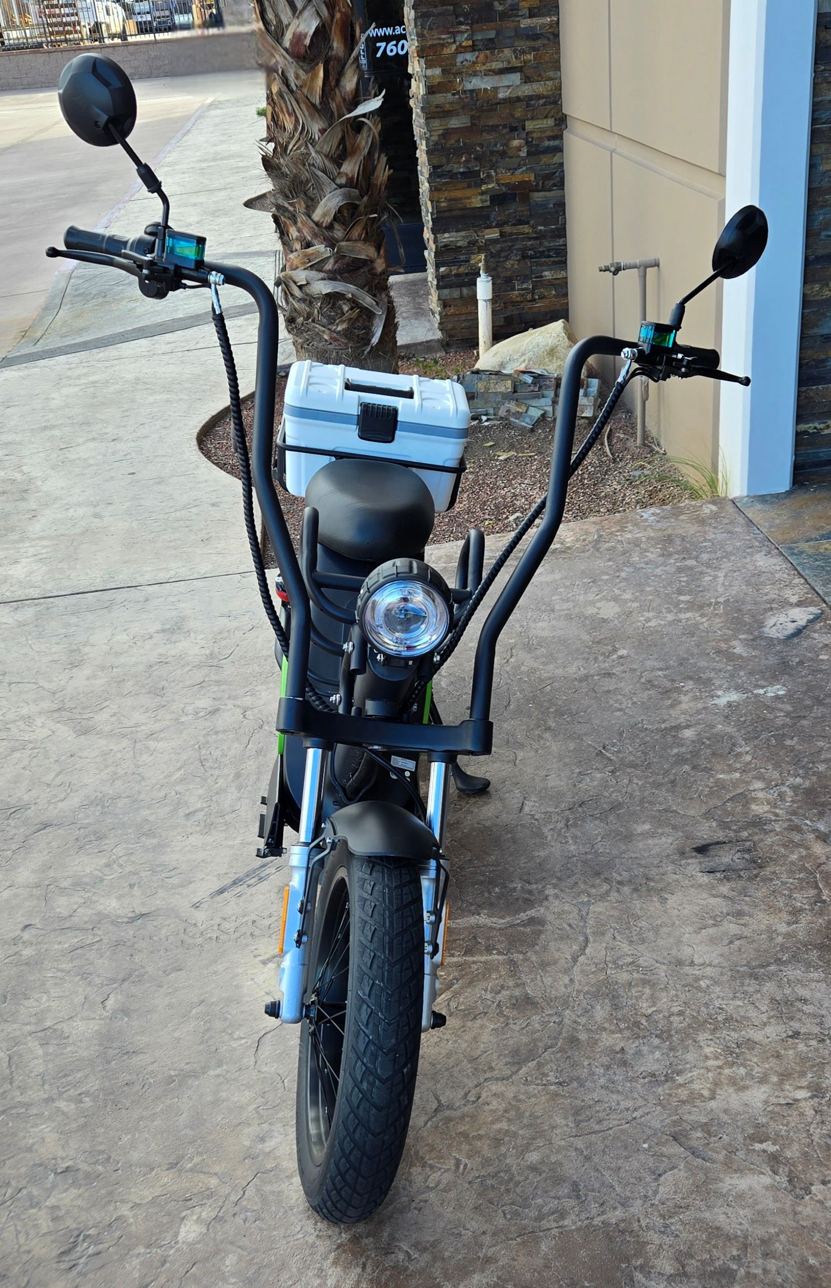 Black and green electric scooter with white storage box, parked on a sidewalk.