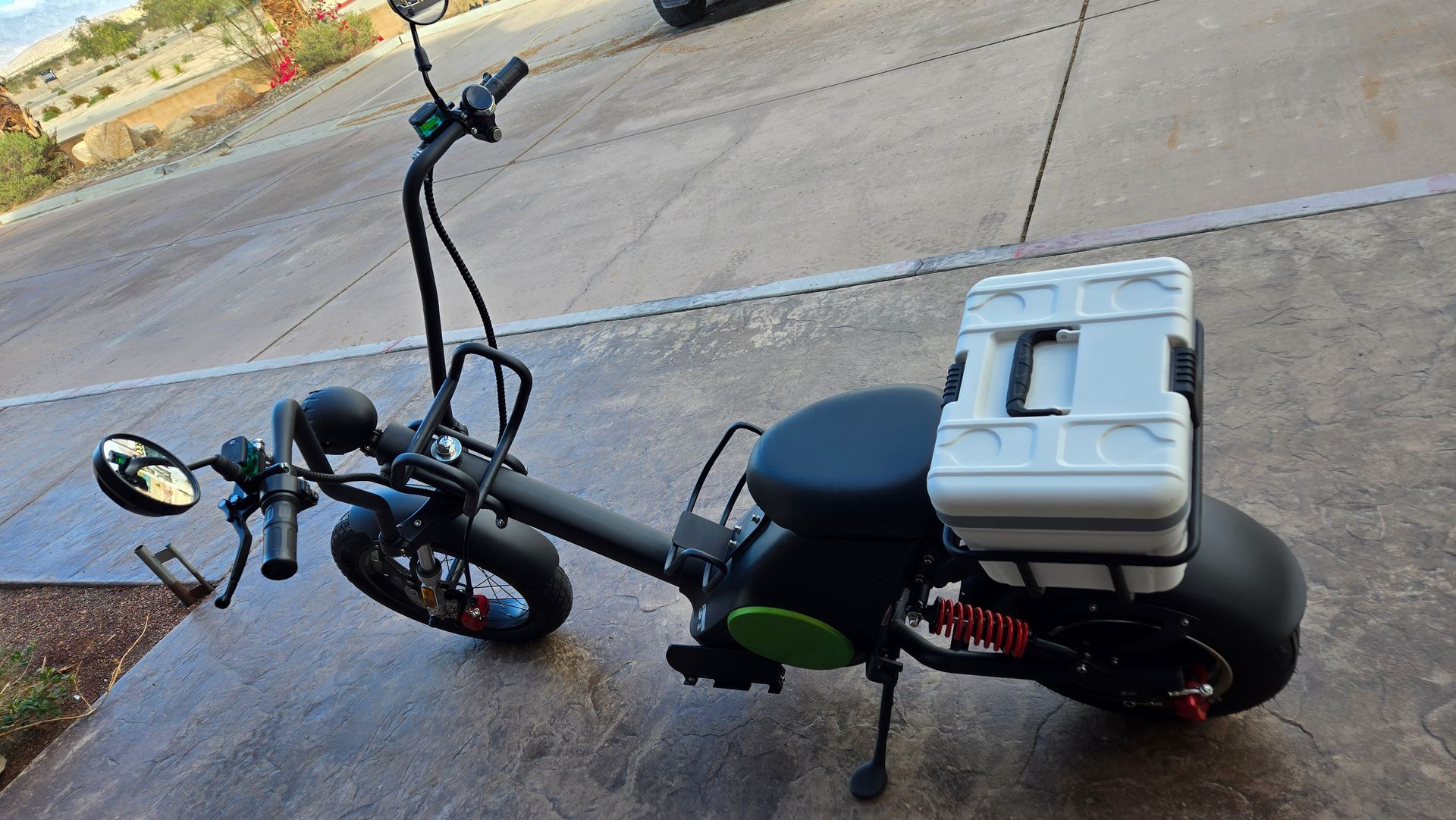 Black electric scooter with white storage box, parked on pavement.