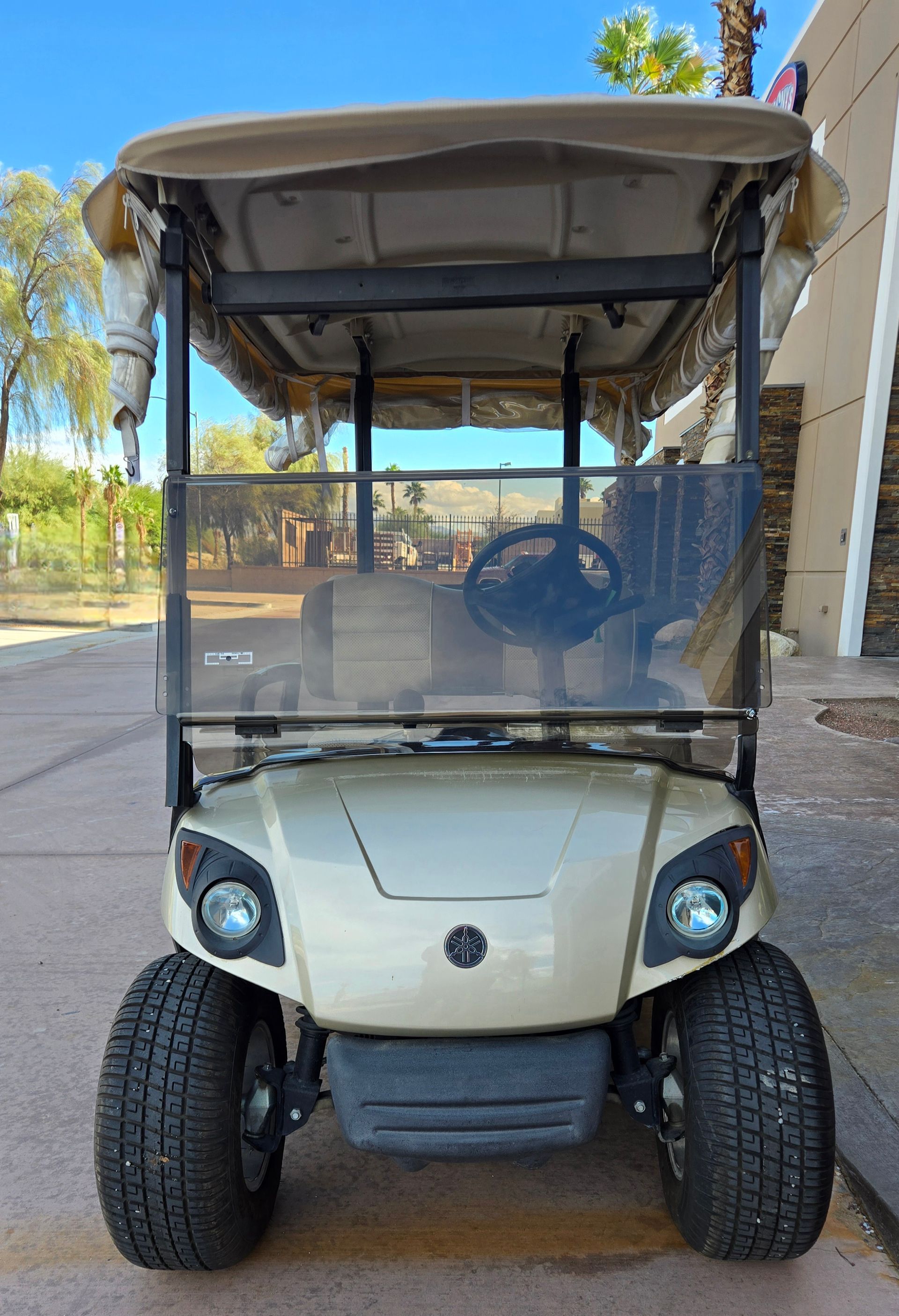 Tan golf cart with windshield and canopy, parked outdoors.