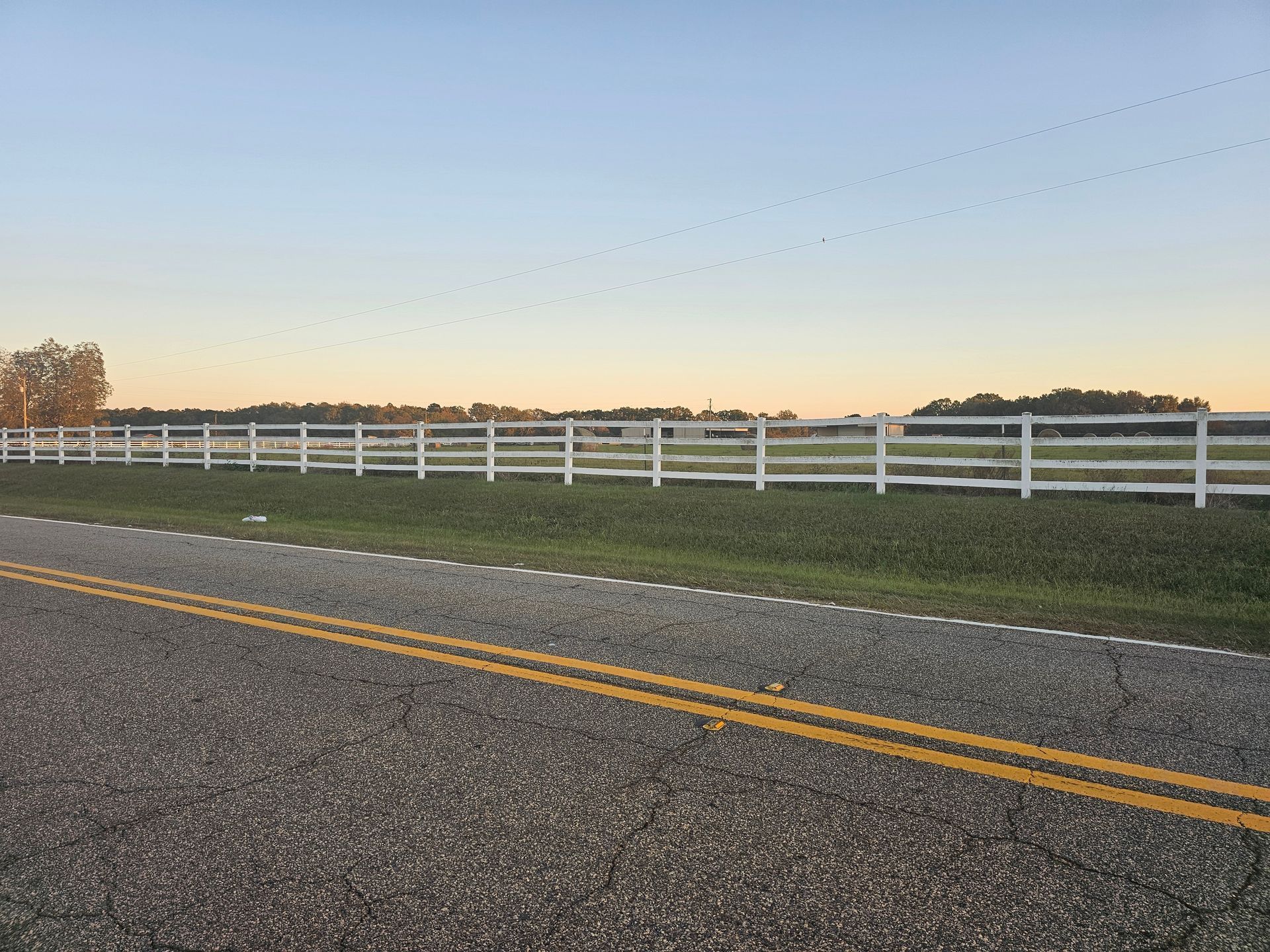 White fence along a road, trees in the background, blue and orange sky.
