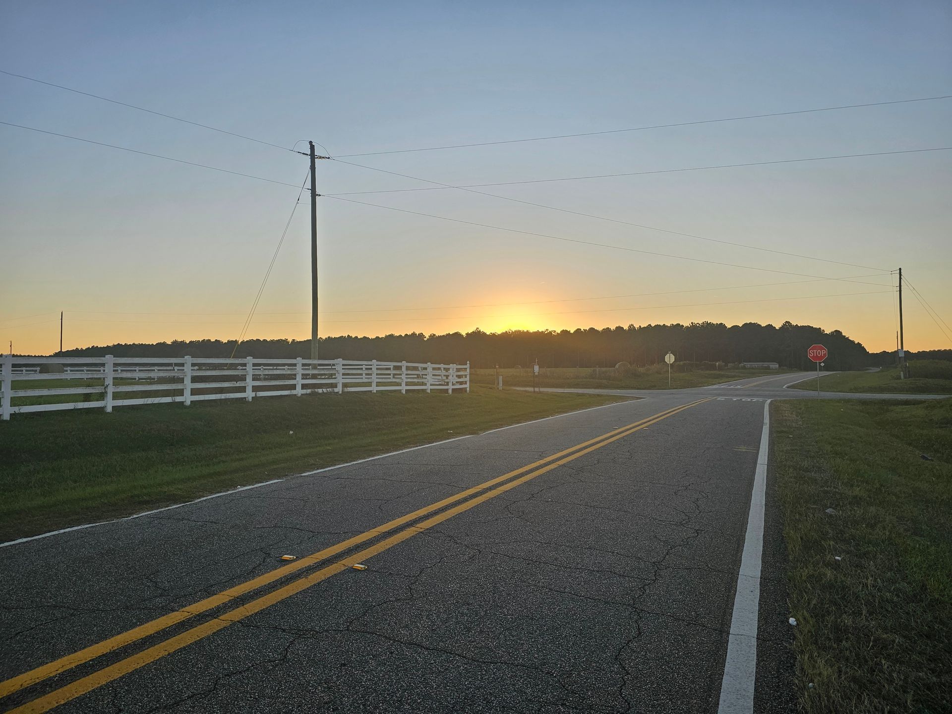 Road at sunset with white fence and trees.