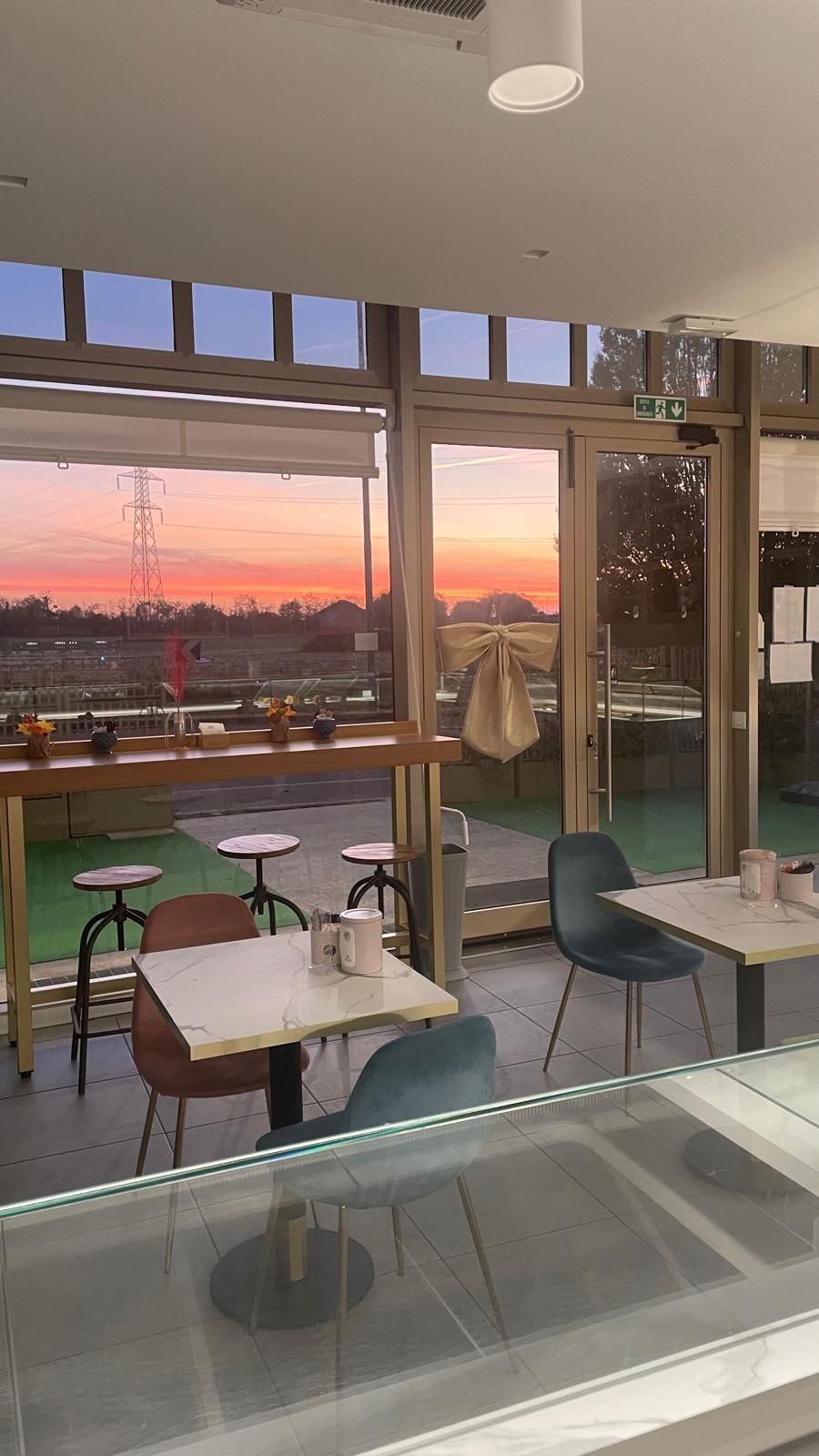 Interno del bar con vista sul tramonto attraverso le ampie vetrate. Tavoli e sedie sono all'interno.