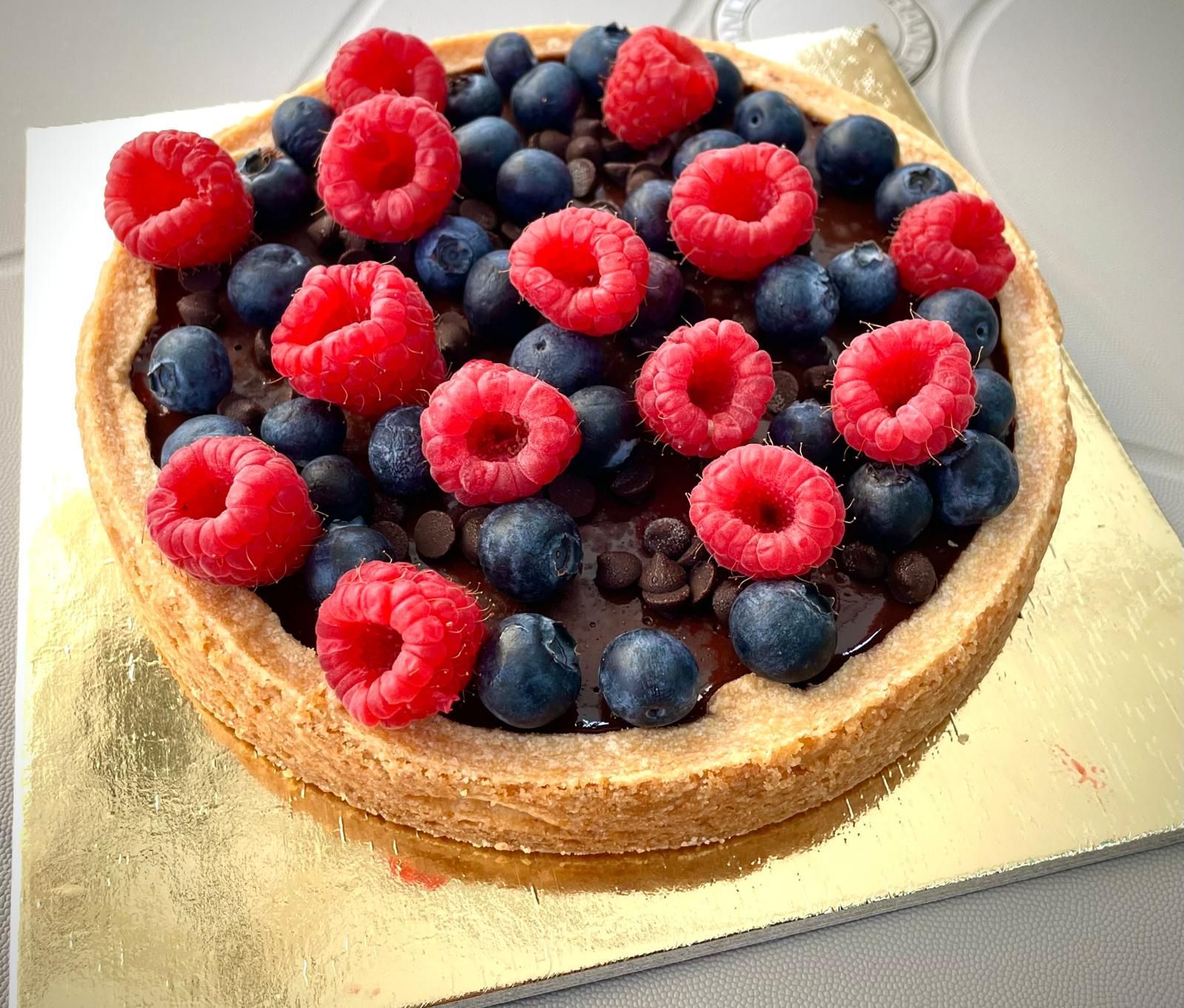 Crostata con crosta dorata, ripieno di cioccolato e guarnita con lamponi e mirtilli freschi su una superficie dorata.