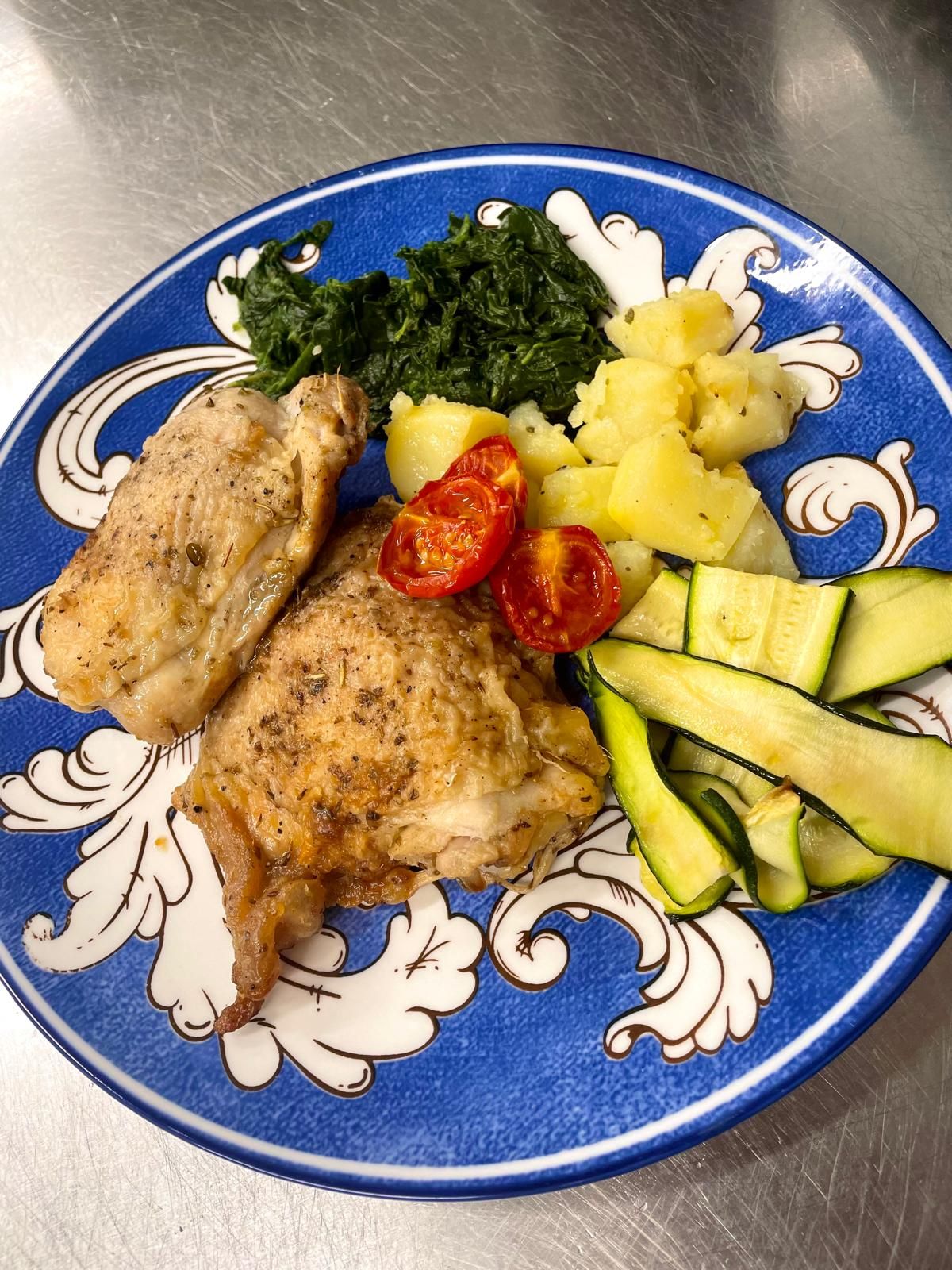 Cosce di pollo, spinaci, patate, pomodori e zucchine su un piatto blu.