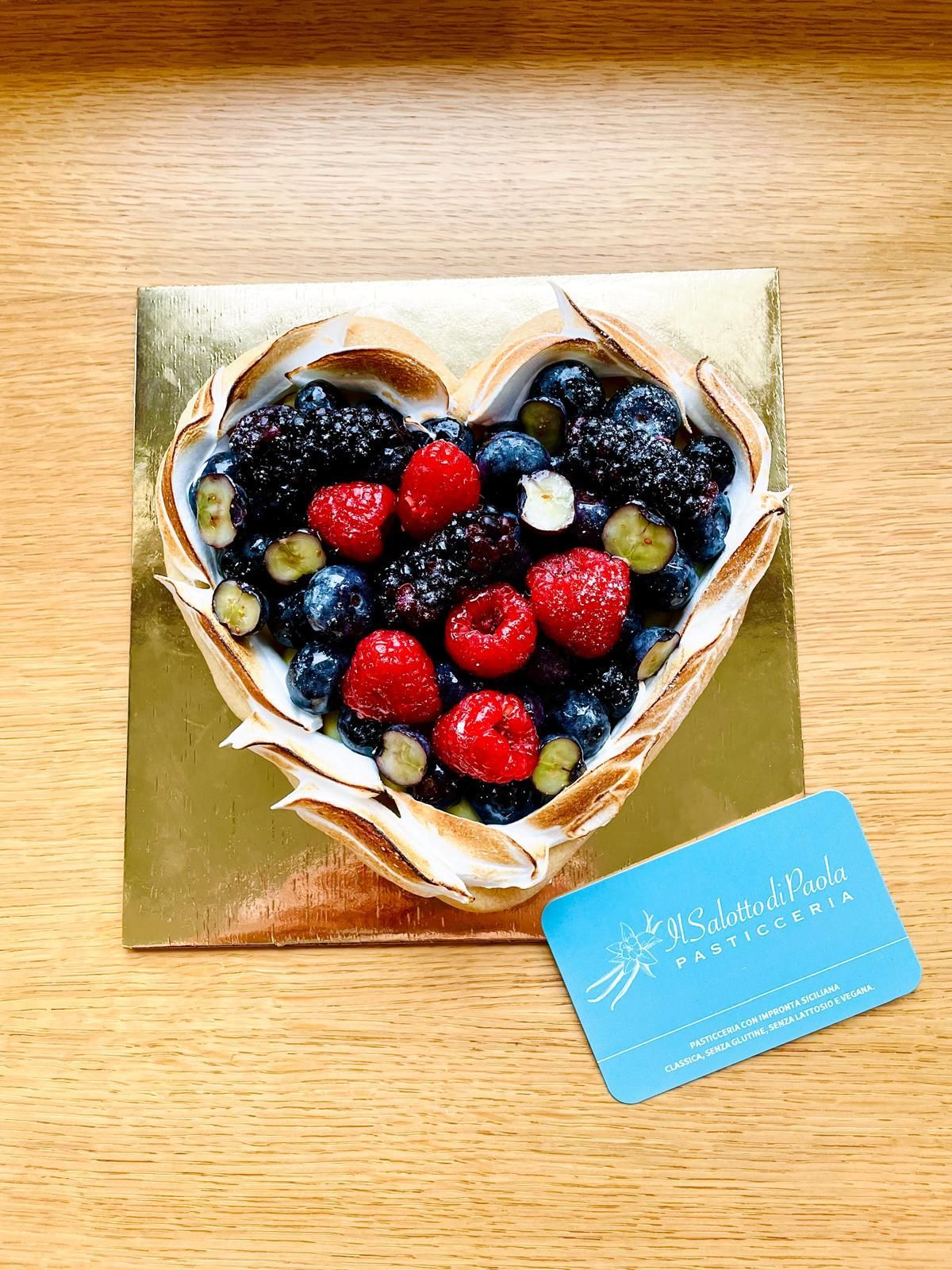 Crostata di frutti di bosco a forma di cuore con base di meringa su una base quadrata dorata e un cartoncino verde acqua.