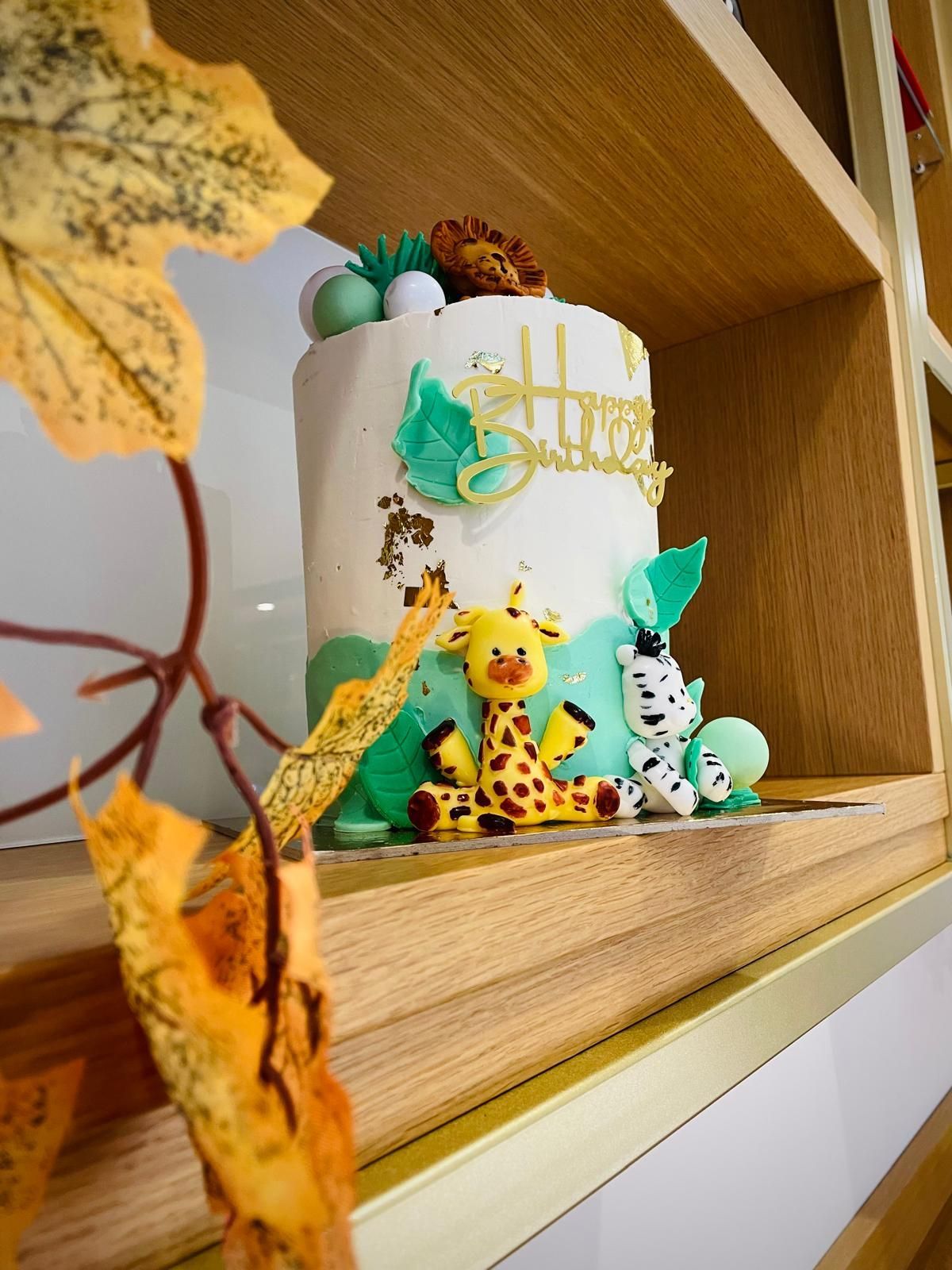 Torta bianca con decorazioni a forma di animali della giungla, dettagli verdi e dorati. Appoggiata su una mensola di legno con foglie autunnali in primo piano.