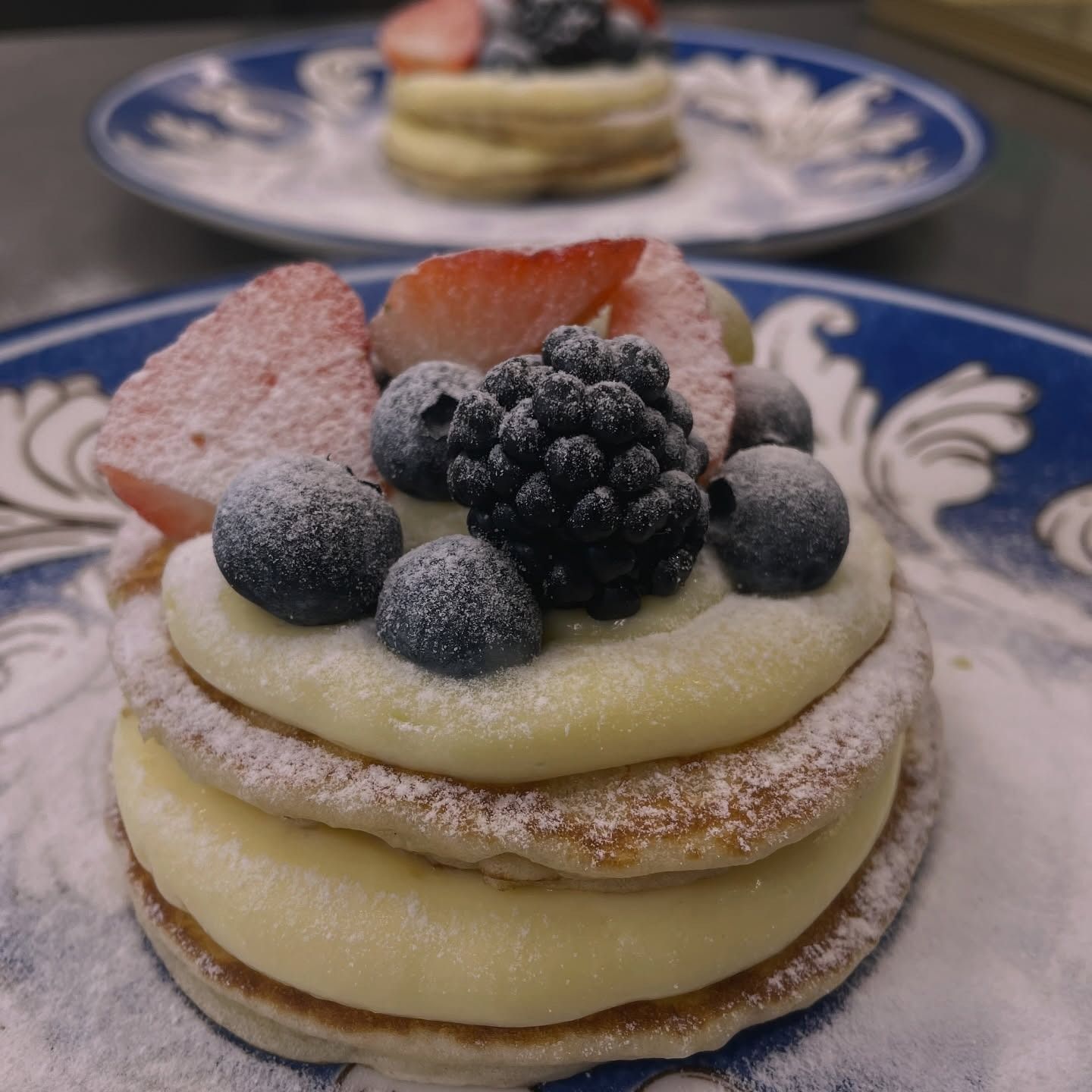 Due pancake impilati con panna, frutti di bosco e zucchero a velo su piatti decorati di blu.