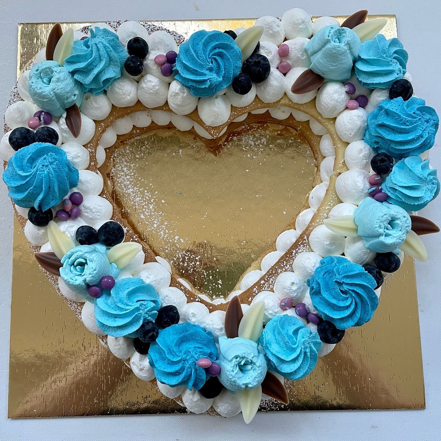 Torta a forma di cuore con fiori di glassa blu, mirtilli e glassa bianca su una superficie dorata.